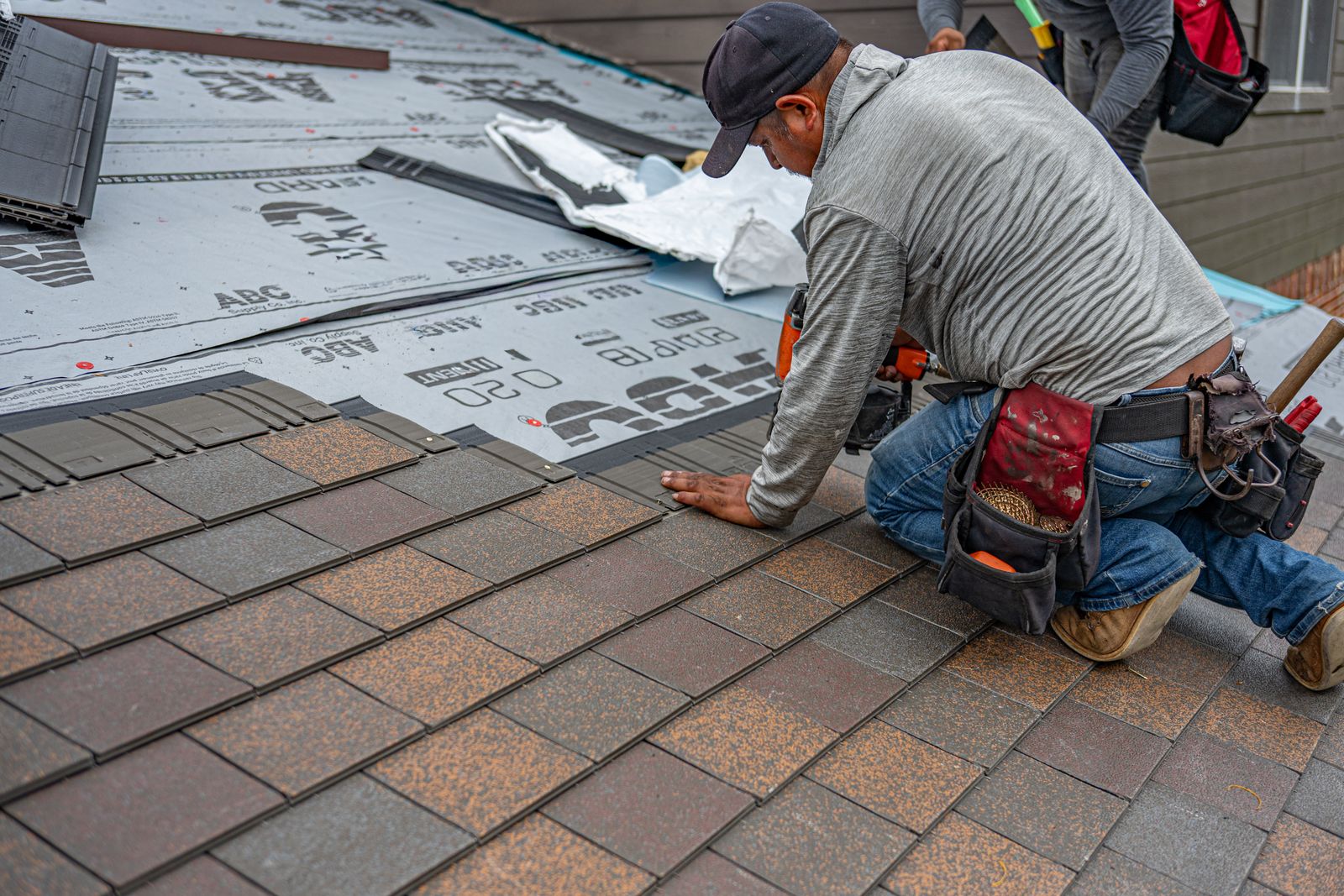 Brown Roofing crew member nailing asphalt shingles over fresh underlayment
